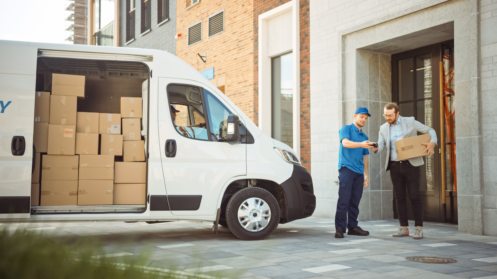 Delivery Man Gives Postal Package to a Business Customer, Who Signs Electronic Signature POD Device. In Stylish Modern Urban Office Area Courier Delivers Cardboard Box Parcel to a Man